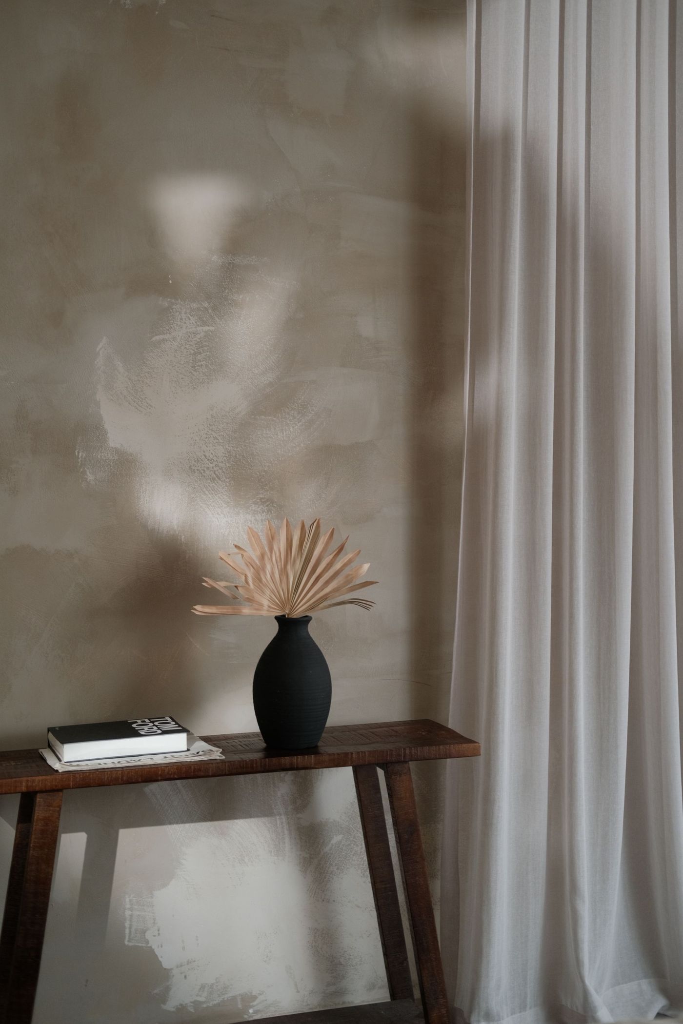 Black vase with dried palm leaves and two books on a dark wooden console, soft light on a plaster wall